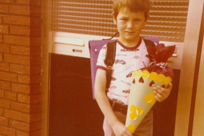 Ein 6-jähriger Junge mit Schultüte steht vor einem Hauseingang eines Mehrfamilienhauses.