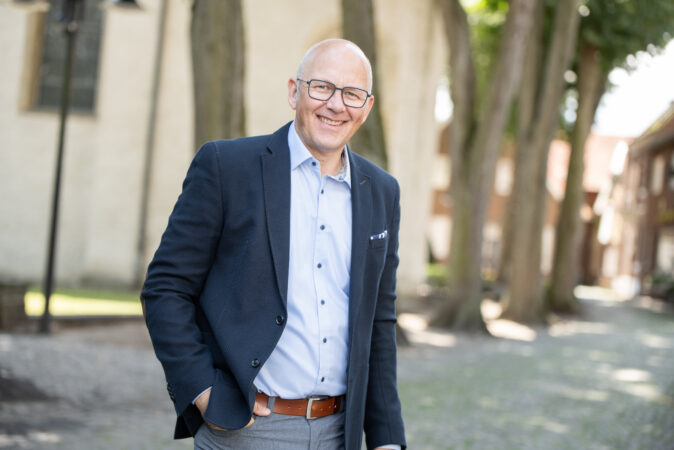 Ein Mann in blauem Sacko und hellblauem Hemd steht vor der Metelener Kirche.
