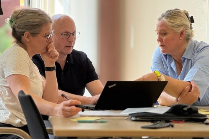 Drei Personen sitzen an einem Tisch und führen ein Gespräch, während sie gemeinsam auf einen Laptop schauen.