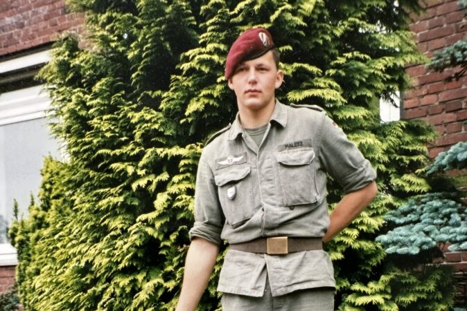 Ein junger Mann mit Fallschirmjägerbarett und 90-er Jahre Bundeswehruniform steht vor einer Zypresse.