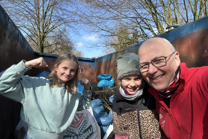 Ein Mann und zwei Kinder lächeln in die Kamera bei sonnigem Wetter, umgeben von Bäumen. Alle wirken fröhlich und entspannt.