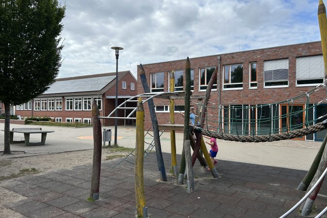 Zwei Kinder spielen auf einem Klettergerüst auf einem Schulhof mit roten Backsteingebäuden im Hintergrund. Der Platz ist gepflastert, daneben stehen ein Baum und ein Tischtennistisch.