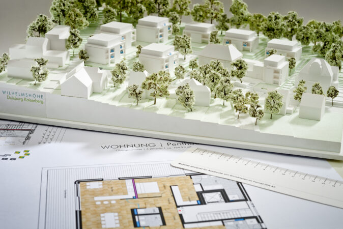 Architekturmodell einer geplanten Wohnsiedlung mit vielen Bäumen und Gebäuden, daneben ein Grundrissplan einer Wohnung. Beschriftung ‚Wilhelmshöhe Duisburg Kaiserberg‘ sichtbar.