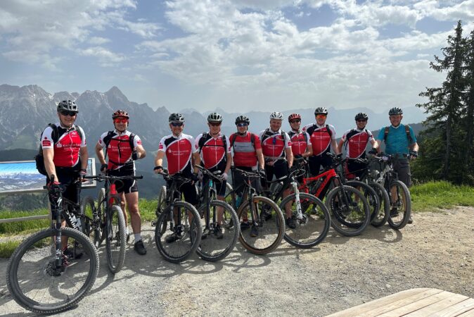Eine Gruppe von 10 Mountainbikern steht nebeneinander aufgereiht auf dem Gibfel eines Berges.