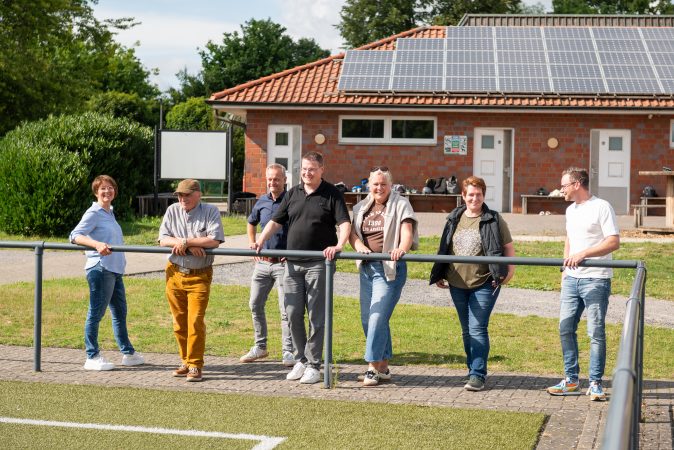 Sieben Erwachsene stehen entspannt an einem Fußballplatz-Geländer und schauen in Richtung Spielfeld. Im Hintergrund ist ein Gebäude mit Solaranlage auf dem Dach zu sehen.